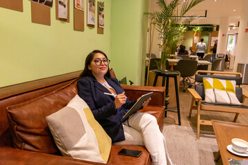 Portrait of young white woman in a busy modern workplace working and  holding phone or tablet in a cowering space