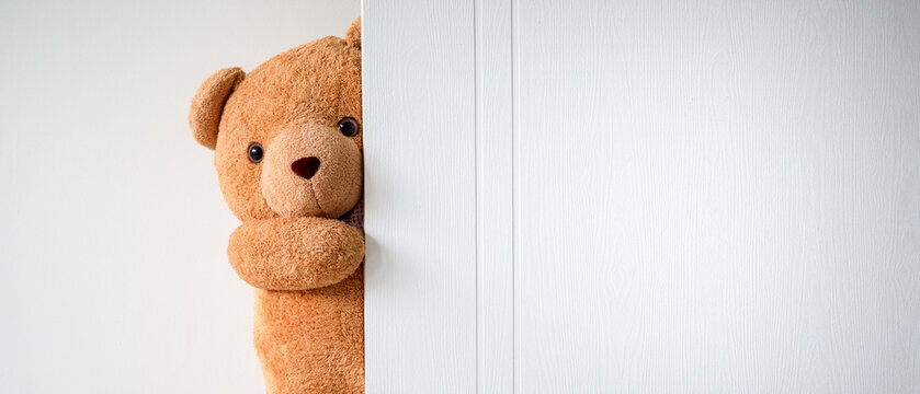 Cute brown teddy bear is hiding behind a white wooden door. Children play with fun and surprises. Copy space for text and content.