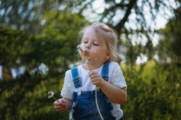 Pretty little girl blowing off dandelion seeds on sunset in summer park. Image with selective focus