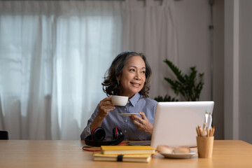 Happy joyful mature laptop user asian woman chatting online, typing, making video call over cup of coffee, tea, relaxing on couch. Senior freelance business lady enjoying working from home