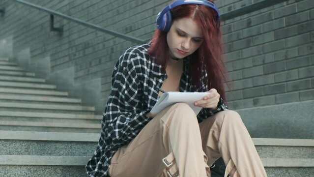 Young attractive girl in casual clothes with red hair is sitting on stairs with headphones and writing in notebook. music college student creates song. creativity and education