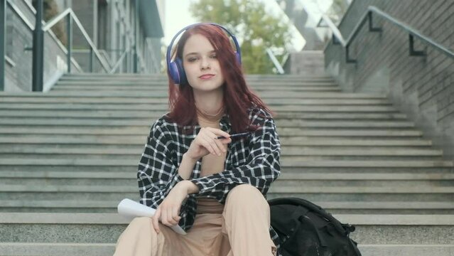 Young attractive girl with red hair sits thoughtfully on stairs with headphones and writes in notebook. student is engaged in creativity to music. creativity and education. teenager in creative search