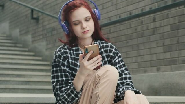 Young attractive Caucasian girl with long red hair sits thoughtfully on street staircase outside alone. Teen hipster puts on headphones and turns on music in modern phone and enjoys audio