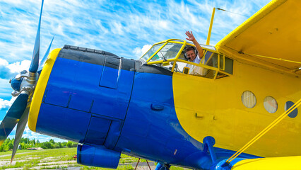Joyful pilot smiles and waves his hand from the cockpit window of an airplane - a biplane. A...