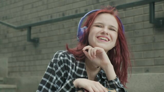 Portrait of an attractive cute girl with long red hair in city. teenage hipster female in headphones listens to her favorite music and laughs looking at camera