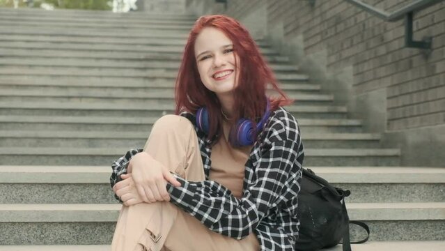 Portrait of joyful carefree girl student with long red hair sitting in city on stone staircase. Happy teen hipster female with headphones listening to favorite music online audio audio enjoying song