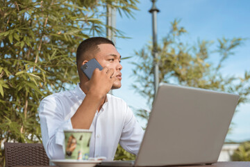 A young entrepreneur contacts a client on his phone while working on some spreadsheets on his...