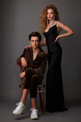 A guy in a brown suit sitting on a chair and a girl in a black long dress posing in the studio on a gray background