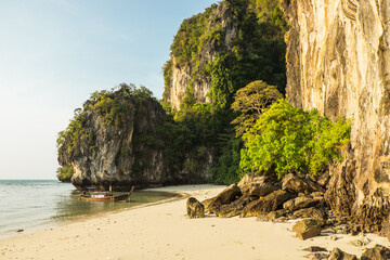 Beach among high cliffs covered with trees on a tropical island, with long tail traditional Thai boat near the shore