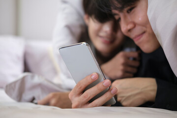 Young gay couple lying under blanket on bed and using smart phone together.