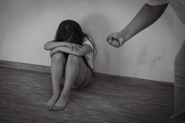 Black and white portrait of little afraid girl sitting with knees up, crossed arms, covering face. Man stretching hand with fist to beat. Protest against domestic violence, abuse, fighting, conflict.