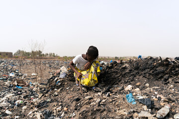Little girl crouching down with a stick in her hand trying to find recyclable material from a...