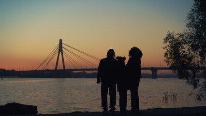 Parents kid silhouette watching sunset standing together at river bridge view.