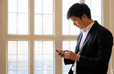 Smiling young asian man using smartphone while standing against wall.