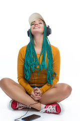 Winsome Smiling Female with African American Dreadlocks Listens Music in Headphones On Cellphone Smartphone While Posing in Streetwear Clothing Over White.
