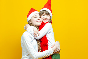 Family of Two Positive Caucasian Girls Sisters  In Festive Santa Hats Having Fun While Embracing And Toching by Cheeks On Yellow Background.