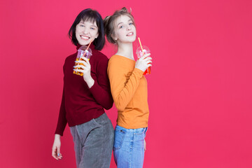 Pair of Winsome Positive Smiling Caucasian Girls Together with Cups Filled With Colorful Juice or Smoothie Drinking While Posing Over Pink Coral Background.