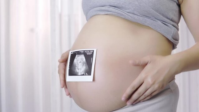 A beautiful pregnant woman is playing with her unborn child, touches her belly with her finger, and gently caresses her stomach. happily In a room with white curtains, slow motion