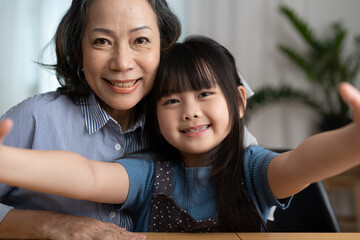 Close-up portrait of joyful happy elderly grandmother is taking selfie with her adorable granddaughter. They are looking at camera with wide smile at home.