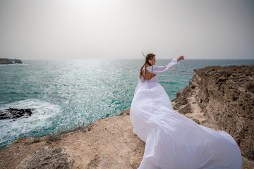 Happy freedom woman on the beach enjoying and posing in white dress. Rear view of a girl in a fluttering white dress in the wind. Holidays, holidays at sea.