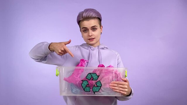 Confident ecofriendly person gen Z with short hair holding container with recycle sign, throwing out plastic garbage into recycling bin, showing how it's done isolated on purple background indoors