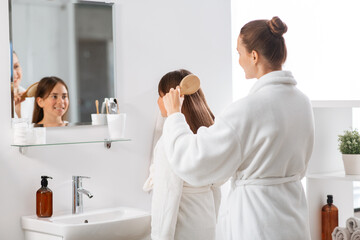 beauty, hygiene, morning and people concept - happy smiling mother and daughter with hairbrush brushing hair at bathroom