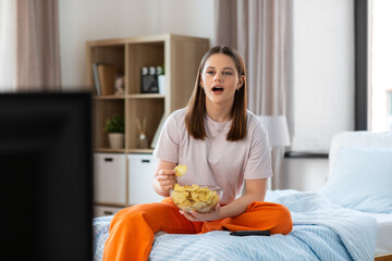 fast food and people concept - happy girl eating crisps and watching tv sitting on bed at home