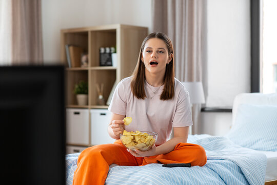 Fast Food And People Concept - Happy Girl Eating Crisps And Watching Tv Sitting On Bed At Home