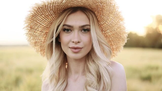 Portrait of attractive blond woman in hat standing in the golden field with beautiful sunset and looking at the camera. 