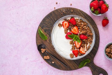 Breakfast, unsweetened yogurt with chocolate baked granola and chopped fresh strawberries in a gray bowl on a pink concrete background. Breakfast recipes.