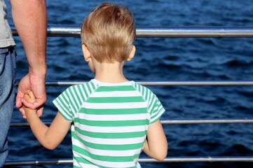 a boy in a striped T-shirt stands by the sea and holds his father's hand rear view selective focus