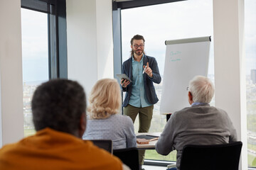 A teacher is teaching a lesson small group of senior students while standing in front of board and holding the tablet.