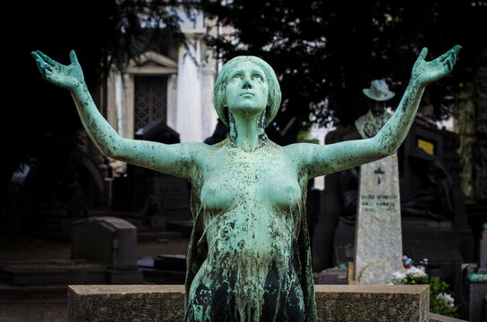 La statua di bronzo di una donna che solleva le braccia al cielo pregando sopra una tomba del cimitero monumentale di Milano