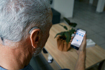 Hearing impaired senior man adjusting settings for his BTE hearing aid via smartphone. Hearing aids with innovative technologies at audiology - obrazy, fototapety, plakaty