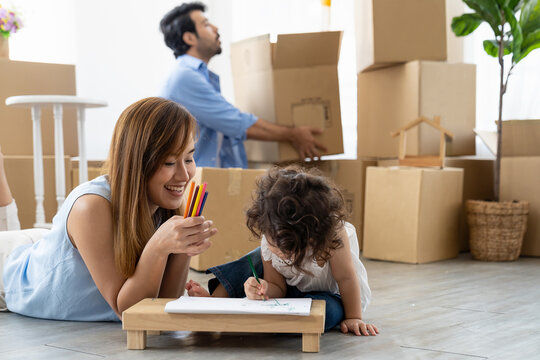 Young mother and her little daughter drawing on pages with colour pencils and man carrying big cardboard box at new home. Family and moving house concept.