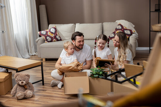 A family unpacks cardboard boxes together after moving into a new apartment. A couple and their children look at old photos in frames, find beautiful keepsakes, tidy up their home.