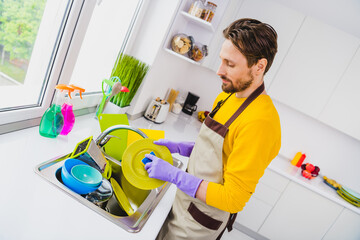 Profile side photo of young handsome man wear apron good mood cleaning wash dishes sink kitchen duty indoors