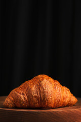 Freshly baked croissant on a wooden podium, dark background. Fresh pastries for French breakfast. Vertical shot, close-up and selective focus on a croissant.