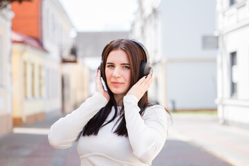 beautiful caucasian young cheerful woman in fashionable clothes listens to music using headphones and a smartphone and smiles while walking around the city