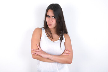 Picture of angry Young beautiful brunette woman wearing white top over white wall crossing arms. Looking at camera with disappointed expression.