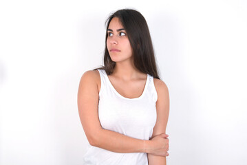 Pleased young beautiful brunette woman wearing white top over white wall keeps hands crossed over chest looks happily aside