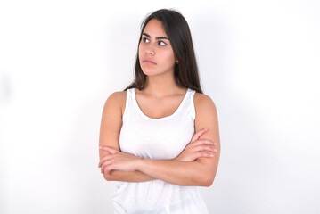 Charming thoughtful young beautiful brunette woman wearing white top over white wall stands with arms folded concentrated somewhere with pensive expression thinks what to do