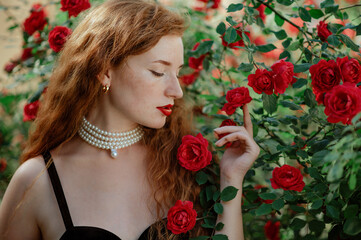 Beautiful redhead freckled woman wearing elegant pearl necklace, golden earrings, posing in red rose garden. Copy, empty space for text - obrazy, fototapety, plakaty