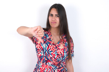 young beautiful brunette woman wearing colourful dress over white wall looking unhappy and angry...