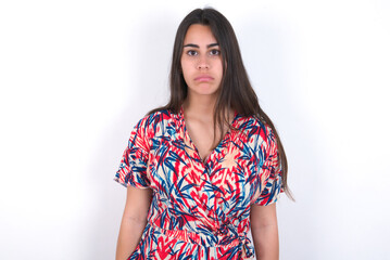 Dissatisfied young beautiful brunette woman wearing colourful dress over white wall purses lips and has unhappy expression looks away stands offended. Depressed frustrated model.