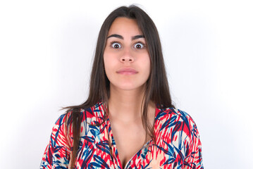 Stunned young beautiful brunette woman wearing colourful dress over white wall stares reacts on...