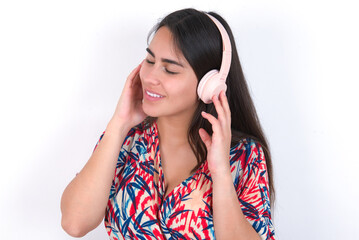 young beautiful brunette woman wearing colourful dress over white wall with headphones on head, listens to music, enjoying favourite song with closed eyes, holding hands on headset.