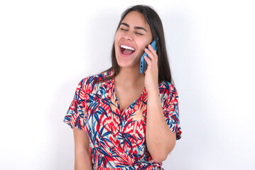 Overemotive happy young beautiful brunette woman wearing colourful dress over white wall laughs out positively hears funny story from friend during telephone conversation