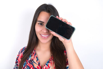young beautiful brunette woman wearing colourful dress over white wall holding modern smartphone covering one eye while smiling