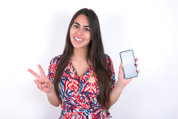 young beautiful brunette woman wearing colourful dress over white wall holding modern device showing v-sign